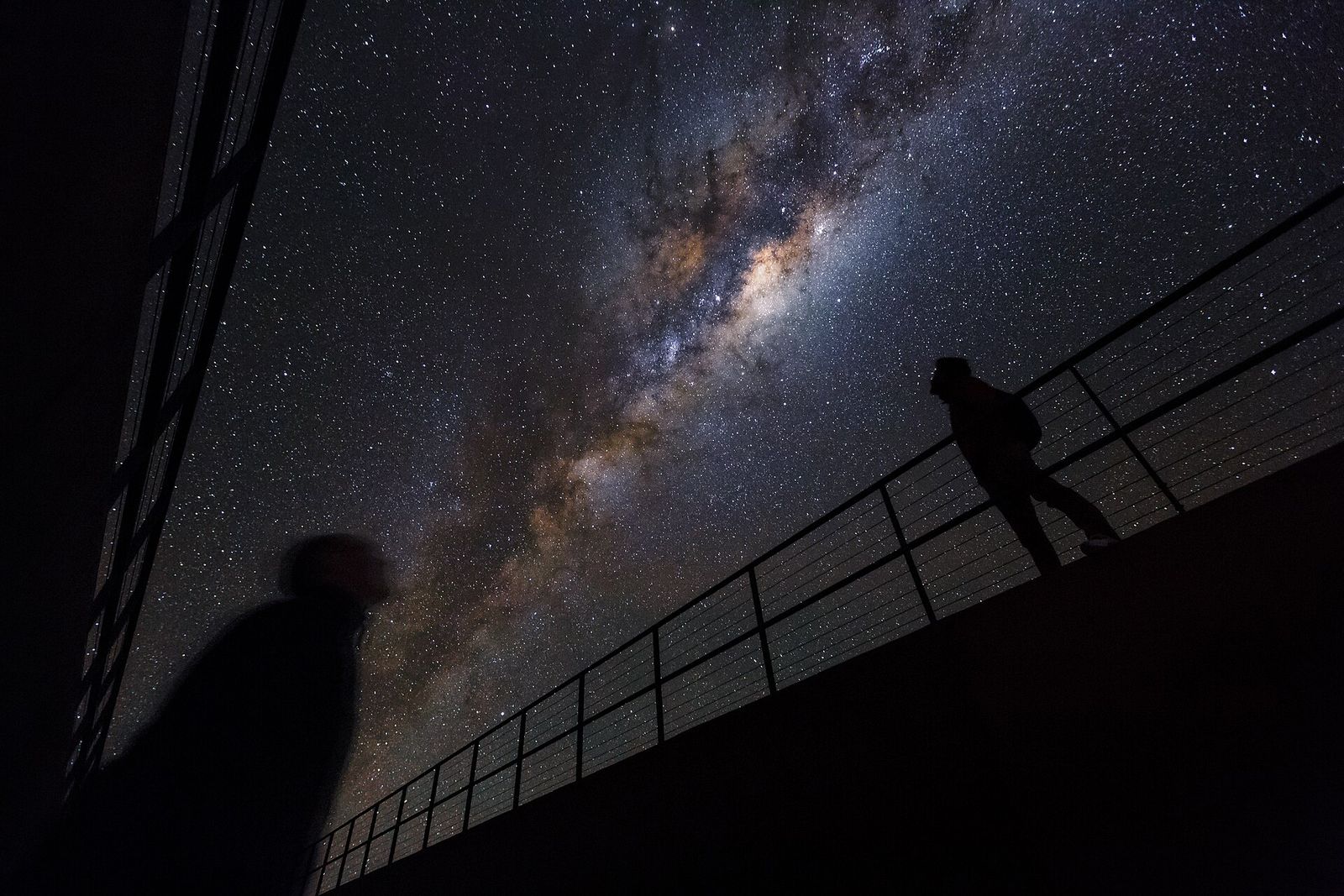 The Milky Way arcing over a walkway railing with two silhouetted stargazers